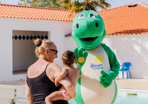 Een vrouw en een kind ontmoeten een groene dinosaurusmascotte bij Flower Camping l'Escale du Perthuis in Frankrijk.
