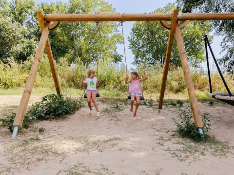 Dos niños se columpian en un parque infantil de arena rodeado de árboles en Flower Camping l'Escale du Perthuis, Pays de la Loire.