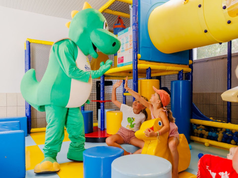 Børn leger med en grøn dinosaurmaskot i et farverigt indendørs legeområde i Flower Camping l'Escale du Perthuis.