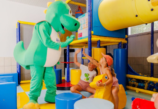 Kinderen spelen met een groene dinosaurusmascotte in een kleurrijke binnenspeelruimte bij Flower Camping l'Escale du Perthuis.
