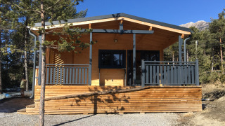 Wooden chalet lodge with air conditioning, surrounded by pines and mountains, sunny day outside.