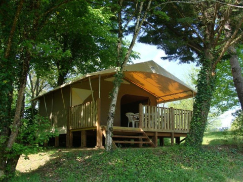 Tenda safari con veranda in legno e sedie di plastica, immersa in una zona boschiva ricca di verde.