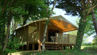 Safari tent with a wooden porch and plastic chairs in a forested area, surrounded by lush green trees.