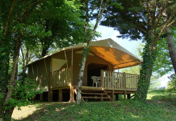 Tenda safari con veranda in legno e sedie di plastica, immersa in una zona boschiva ricca di verde.
