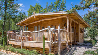 Een rustiek houten chalet met veranda, gelegen in het groen tussen bomen en planten bij een blauwe lucht.