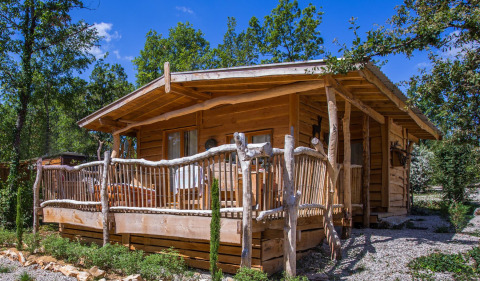 Uno chalet di legno rustico con veranda, immerso tra alberi e vegetazione sotto un cielo azzurro limpido.