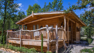 A rustic wooden chalet lodge with a porch, set amongst trees and greenery under a bright blue sky.