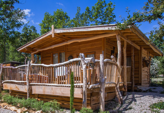 Uno chalet di legno rustico con veranda, immerso tra alberi e vegetazione sotto un cielo azzurro limpido.