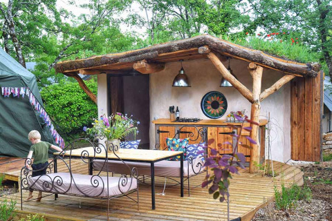 Cucina all'aperto e terrazza con panchine accanto alla tenda safari Dôme immersa nel verde della natura.