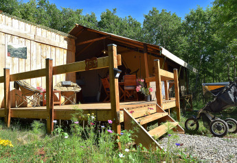 Tente safari avec terrasse privée et sanitaires, entourée de verdure, avec poussette au premier plan.