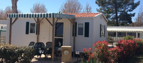 Et foto af en mobilhome med en terrasse, udendørs borde og stole, omgivet af buske og træer.