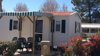 Foto eines Mobilheims mit überdachter Terrasse, Gartenmöbeln und umgeben von blühenden Büschen.