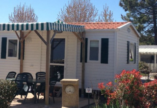 Photo d’un mobil-home avec terrasse couverte, table et chaises extérieures, entouré de verdure.