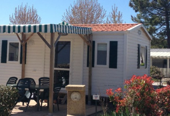 Photo d’un mobil-home avec terrasse couverte, table et chaises extérieures, entouré de verdure.