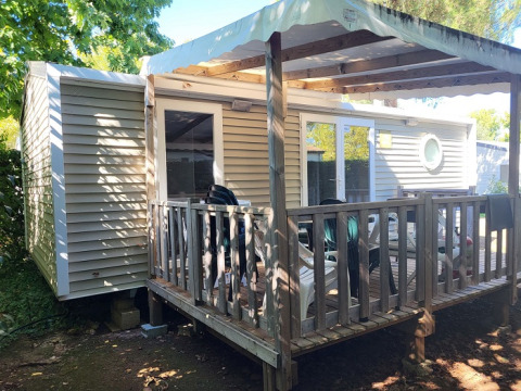 Casa mobile con veranda in legno e tettoia bianca, circondata da alberi e sedie sulla terrazza.