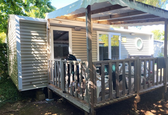 Casa móvil con porche de madera y toldo blanco, rodeada de árboles y sillas en la terraza exterior.
