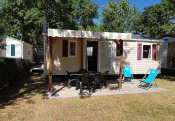 Casa mobile con veranda coperta, sedie da giardino e sdraio, circondata da alberi e prato.