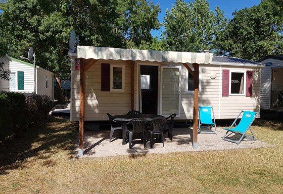 Mobil-home avec terrasse couverte, chaises de jardin et transats, entouré d’arbres et de pelouse.