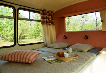 Interieur van een knusse schoolbuslodge met bed, grote ramen en uitzicht op groen bij Les 3 Cantons.