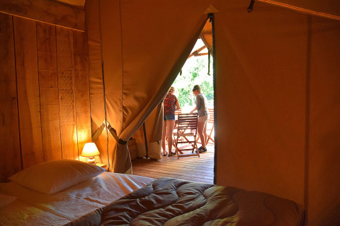 Interieur van een safaritent met bed en lamp, uitkijkend op terras met twee mensen buiten.