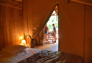Interieur van een safaritent met bed en lamp, uitkijkend op terras met twee mensen buiten.