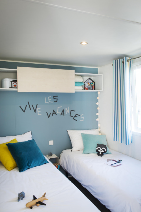 Deux lits simples dans une chambre lumineuse, mur bleu, coussins, jouets et inscription 'Les Vive Vacances'.