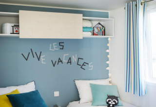 Deux lits simples dans une chambre lumineuse, mur bleu, coussins, jouets et inscription 'Les Vive Vacances'.