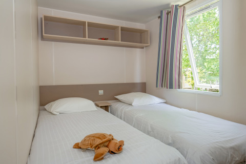 Schlafzimmer mit zwei Einzelbetten, weißer Bettwäsche, Stofftier und Fenster im Mobile home «Midi Pyrénées».