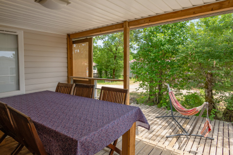 Covered terrace at Mobile home “Midi Pyrénées” at Campsite Les 3 Cantons with dining table and hammock.