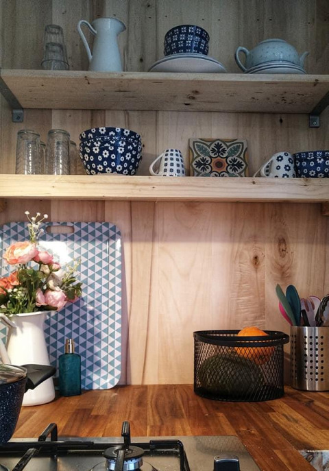 Acogedora cocina pequeña con estantes de madera, vajilla azul, cesta de frutas y jarra con flores.