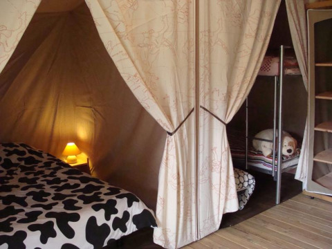 Interieur van een safaritent met tweepersoonsbed, stapelbedden, warme lamp en open opbergrekken.