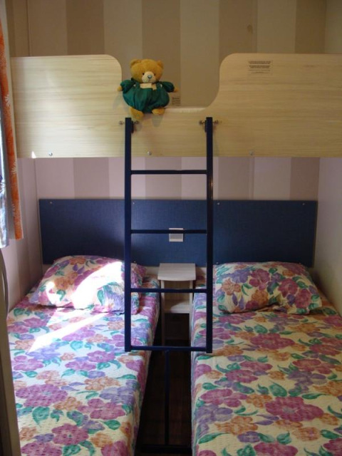 Chambre dans une mobil-home avec lit superposé, deux lits jumeaux fleuris et un ours en peluche en haut.