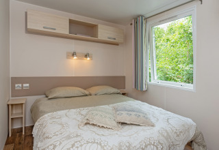 Schlafzimmer in Mobile Home «Loggia» auf dem Campingplatz Les 3 Cantons, Frankreich, mit Doppelbett und Fenster.