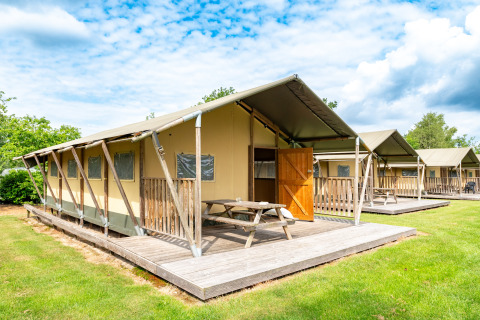 Tienda safari Woodlodge con terraza de madera, mesa de picnic y puerta abierta sobre césped y cielo azul.
