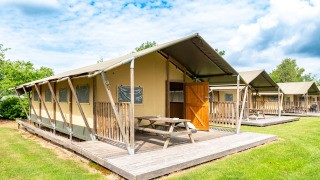 Tienda safari Woodlodge con terraza de madera, mesa de picnic y puerta abierta sobre césped y cielo azul.