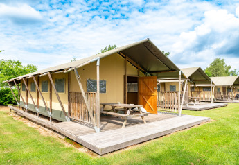 Tenda safari Woodlodge con veranda in legno, tavolo da picnic e porta aperta su prato verde e cielo azzurro.