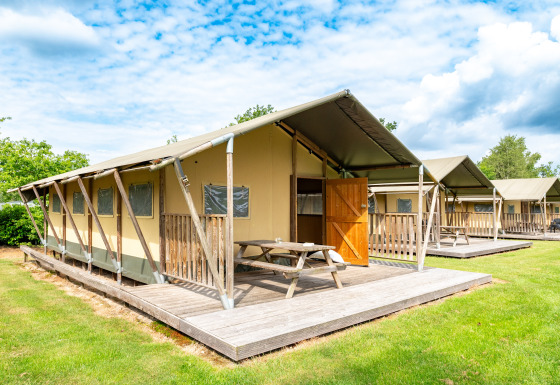 Tente safari Woodlodge avec terrasse en bois, table de pique-nique et porte ouverte, sous ciel bleu nuageux.