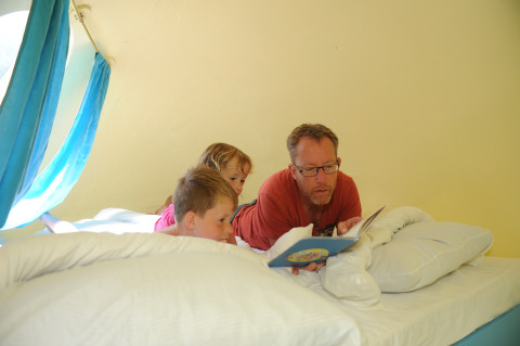 A man reads a book to two children on a bed inside a cozy pod at Veluwe campsite 't Schinkel in the Netherlands.