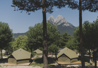 Tiendas safari en un camping rodeadas de árboles y una montaña imponente como fondo bajo el cielo azul.