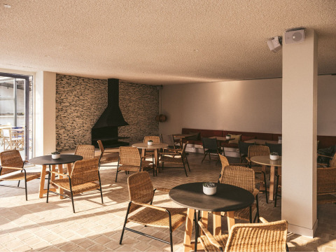 Bright lounge area at Wecamp Cadaqués, Catalonia, with wicker chairs, round tables, and a stone fireplace.