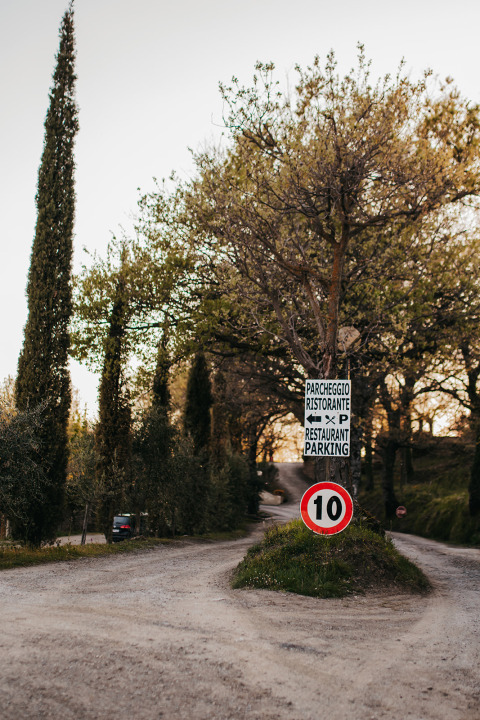 Entrance to Campsite Lucherino in Tuscany, Italy, with gravel road, trees, and signs for restaurant and parking.