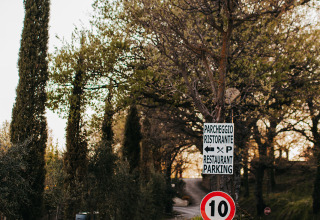 Entrée du camping Lucherino en Toscane, Italie, avec chemin de gravier, arbres et panneaux vers restaurant et parking.