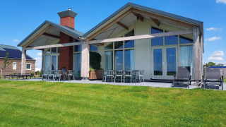 Alojamiento glamping moderno con grandes ventanales, patio y césped bajo un cielo azul y nubes blancas.