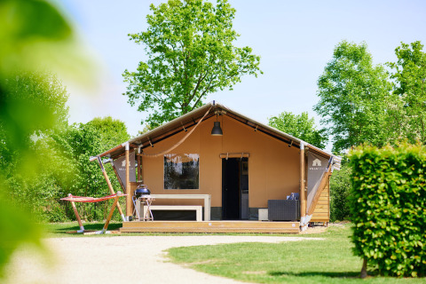 Safaritent Nomad met veranda, barbecue en hangmat, omringd door groene bomen en zonlicht.