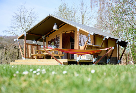 La tenda safari Compact si trova su un prato, con amaca, terrazza in legno e tavolo da picnic esterno.
