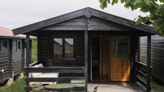 Pequeña casa de madera negra con porche y muebles de jardín verdes sobre césped, bajo cielo nublado.
