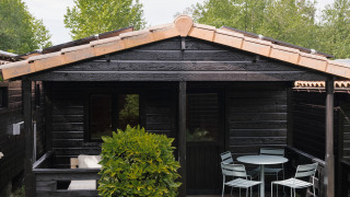 Bungaló de madera negra con terraza pequeña, muebles de jardín y vegetación en Bungalow Basic Family Plus.