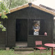 Een vrouw met een kind staat in de deuropening van een zwart houten huisje genaamd Bungalow Basic Couple in de regen.