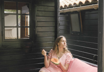 Woman in a pink dress eating melon on a striped sofa in the Basic Couple bungalow at Wecamp Cudillero, Spain.