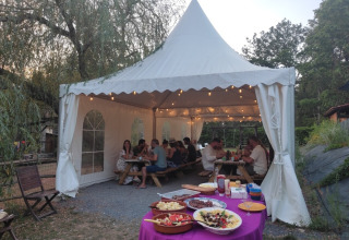 Cena all’aperto sotto un tendone al Campeggio Pagel, Occitania, Francia, con cibo e ospiti seduti.